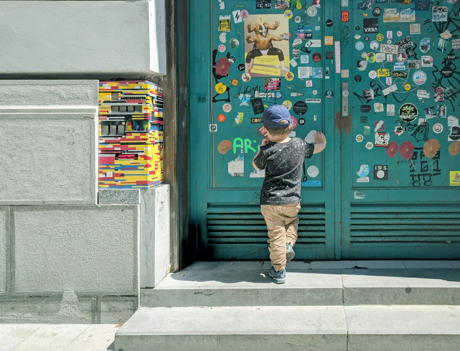 Child exploring a colorful doorway adorned with stickers and LEGO art in a city setting.