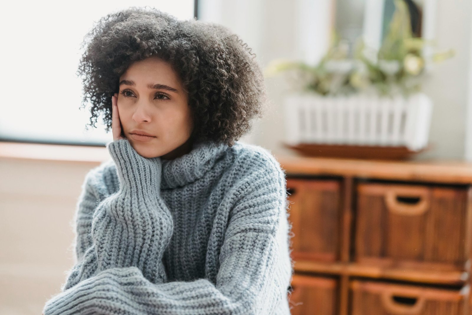 Discontent African American female leaning on hand and looking away while sitting in light room near green plant on blurred background