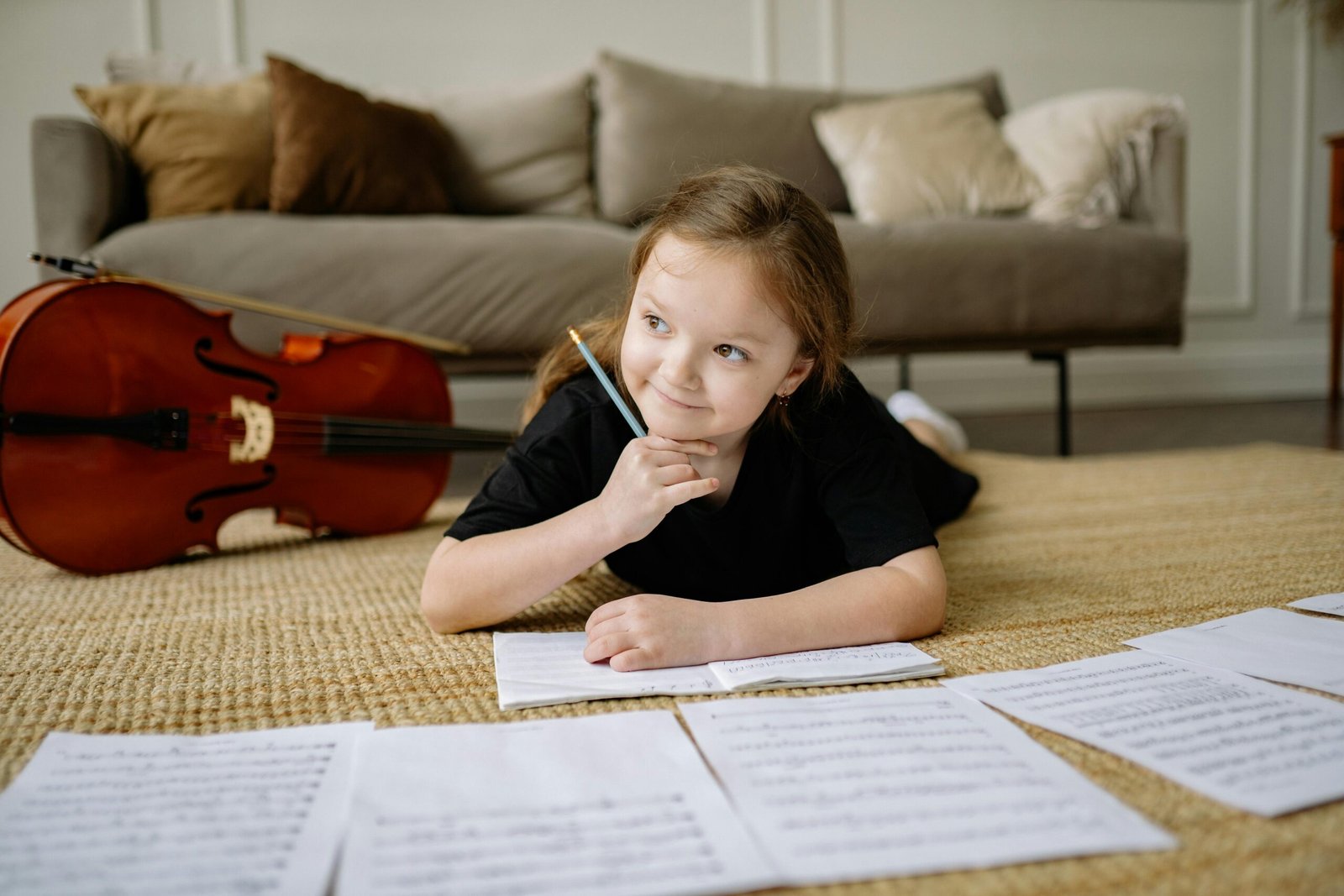 A girl lying on the carpet, pondering over music sheets with a cello beside her.